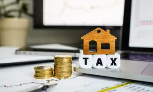 Drewniany domek na kostkach z napisem TAX, obok stosy monet. Symbolizuje koszty związane z nieruchomościami, w tym pcc.