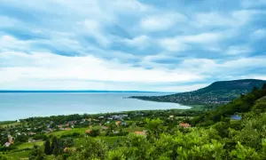 Widok na jezioro Balaton z zielonymi wzgórzami i domkami. Chmury na niebie tworzą dramatyczny krajobraz.