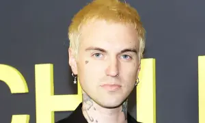 A young man with blonde hair, blue eyes, and tattoos, including "AUTOPILOT" on his neck, poses for a photo. He wears hoop earrings and a nose ring. His style is reminiscent of jordan jutes.