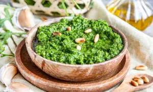 Frisches Rucola Pesto in Holzschale, garniert mit Pinienkernen. Daneben Knoblauch und Olivenöl.