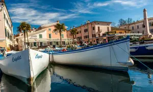 Barche a Cisano di Bardolino, con palme e case colorate sullo sfondo. Il sole splende su un cielo azzurro e limpido.
