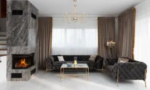 Elegancki salon z kominkiem, ciemnymi sofami i złotymi akcentami. Zasłony w kolorze kość słoniowa dodają ciepła.