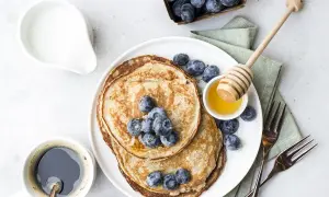 Pancake senza uova con yogurt, guarniti con mirtilli freschi e miele. Un caffè nero e una brocca di latte completano questa deliziosa colazione.