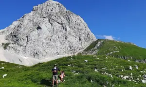 Rodzina w górach: mężczyzna z dzieckiem w nosidle i dziewczynka na tle skalistego szczytu i zielonych zboczy.