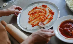 Ręce rozsmarowują pyszny sos pomidorowy do pizzy na cieście. Obok miseczka z sosem i tarta mozzarella.