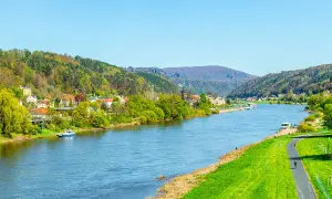 Blick auf den Elberadweg mit Radfahrern und einer Stadt am Flussufer. Die Elbe fließt durch eine grüne Landschaft mit bewaldeten Hügeln. Die Elberadweg Entfernungen sind hier gut zu erkunden.