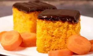 Due fette di torta di carote, soffice e arancione, ricoperte da glassa al cioccolato fondente. Accanto, fette di carota fresca.