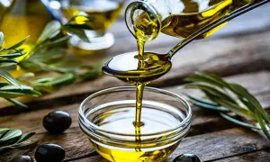 Olio di oliva non extravergine versato da un cucchiaio in una ciotola, con olive e rami d'ulivo sullo sfondo.