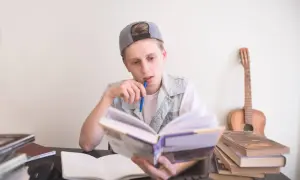 Chłopak w czapce z daszkiem, z długopisem w ustach, pochłonięty czytaniem na czas. Obok gitara i stosy książek.