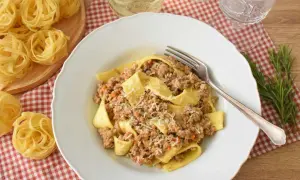 Pappardelle z bogatym ragu, które to danie, posypane parmezanem. Obok surowy makaron i gałązka rozmarynu.
