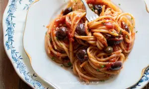 Un piatto invitante di spaghetti alla puttanesca, con olive nere, capperi e un sugo rosso vivace, pronto per essere gustato.