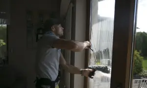 Mężczyzna w czapce z daszkiem pokazuje, jak myć okna ściągaczką, usuwając pianę z szyby.