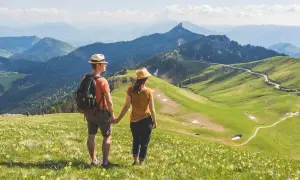 Couple en chapeau admirant un paysage de montagne verdoyant, parsemé de fleurs jaunes. Une rando Chartreuse insolite, avec des sentiers sinueux et des sommets bleutés à l'horizon.
