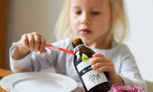 Mała dziewczynka trzyma butelkę oleju z czarnuszki, obok talerz i słoiczek z dżemem.
