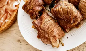 Involtini di carne succulenti, cotti alla perfezione, serviti su un piatto bianco. A lato, fette di prosciutto crudo in un cestino di legno.