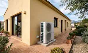 Unidad exterior de bomba de calor aerotermia junto a una casa con fachada amarilla y patio de baldosas.