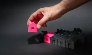 Mano che sceglie una casetta rosa tra tante nere. Un'idea di permuta immobiliare con costruttore.