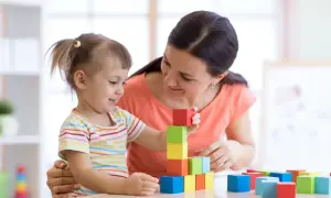 Uśmiechnięta kobieta i dziewczynka budują wieżę z kolorowych klocków. To obrazek, który może inspirować, jak zatrudnić nianię.