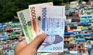 Dłoń trzyma banknoty wonów: 10000, 5000 i 1000. Tło to kolorowe domy, sugerujące pytanie: ile kosztuje wyjazd do Korei Południowej?