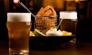 Due bicchieri di birra bionda ghiacciata accompagnano un cestino di anelli di cipolla fritti, patatine e una salsa cremosa.
