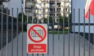 Znak zakazu wjazdu na drogę pożarową na metalowej bramie. W tle budynki mieszkalne i polska flaga.