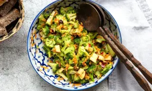 Frischer Brokkoli Salat mit Apfelstücken und Karottenraspeln, serviert in einer blauen Schüssel mit Holzkochlöffeln.