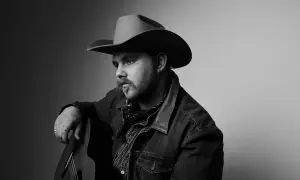 Ernest Keith Smith, a thoughtful country musician, poses with his guitar, wearing a cowboy hat and denim jacket.