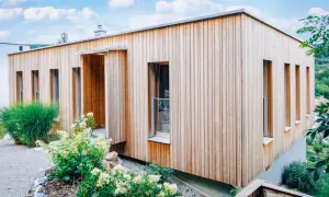 Modernes Haus mit einer Mischfassade aus Holz und Putz. Vertikale Holzverkleidung umrahmt schmale Fenster. Grüne Pflanzen schmücken den Eingangsbereich.