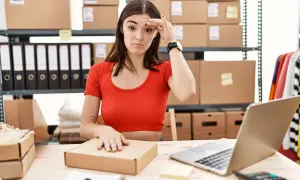 Mujer frustrada revisando el inventario online, rodeada de cajas y carpetas.