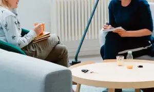 Dwie osoby siedzą w przytulnym pokoju, jedna pije z kubka, druga pisze w notatniku.