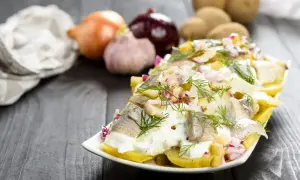 Pyszna sałatka ziemniaczana ze śledziem, udekorowana koperkiem i czerwoną cebulką. Obok leżą ziemniaki, cebula i czosnek.