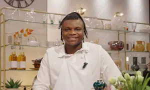Kelvin Wokomaty, en glad kock i vitt förkläde, ler mot kameran. Bakom honom syns hyllor med flaskor och dekorationer.