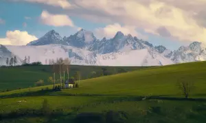 Zielone wzgórza, mały biały domek z drzewami i ośnieżone góry w tle.
