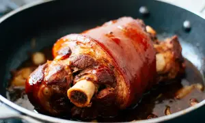 Un magnifique jarret de porc au four, la peau dorée et brillante, mijote dans sa sauce savoureuse avec des gousses d'ail.