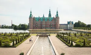 Ett imponerande slott i Danmark, omgivet av vatten och med en vacker barockträdgård. Ett par promenerar längs en damm framför slottet.