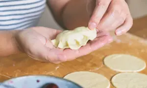 Dłonie formują pieroga z ciasta na pierogi bez jajka. Na blacie leżą okrągłe placki ciasta.