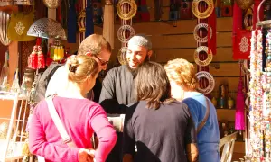 Un groupe de touristes discute avec un vendeur dans un souk de Marrakech, entouré de lanternes, de bijoux et de textiles colorés.