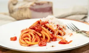 Ein Teller Pasta all'amatriciana, mit Tomaten und geriebenem Käse bestreut. Im Hintergrund ein Stück Guanciale, die Zutat für das authentische amatriciana rezept.
