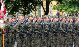 Żołnierze w mundurach defilują z karabinami. W tle sztandar i drzewa. Kategoria e wojsko.