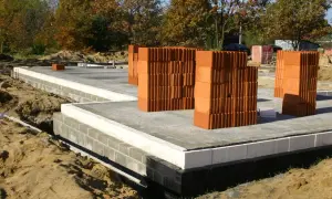 Stosy cegieł, gotowe do budowy domu. Fundament z bloczków i izolacji, piasek wokół. Chudziak czeka na swoją kolej.