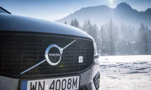 Szary Volvo XC60 Polestar z emblematem Polestar na grillu, zaparkowany na tle ośnieżonych gór i lasu.