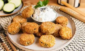 Knusprig frittierte Zucchini-Patties auf einem Teller mit Dip. Daneben liegen frische Zucchinischeiben.