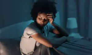 A woman sits in bed, head in hand, eyes closed, appearing to be experiencing fragmented sleep causes.