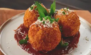 Trois arancini, spécialité italienne, dorés et croustillants, servis sur une sauce rouge avec des herbes fraîches et du parmesan râpé.