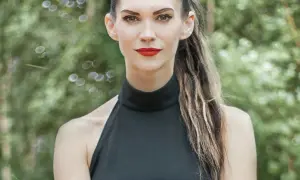 Małgorzata Oliwia Sobczak w czarnej, eleganckiej sukience, z burzą włosów i wyrazistym makijażem, na tle zieleni.