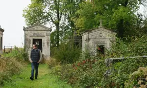 Un homme marche dans un cimetière abandonné en France, entouré de mausolées envahis par la végétation.