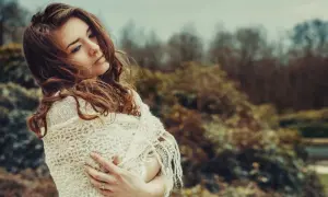 Una giovane donna con capelli castani, avvolta in una sciarpa di pizzo, sembra pensare a se stessa in un parco autunnale.