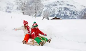 Zwei Kinder rodeln fröhlich durch den Schnee. Ein idyllisches Bild vom Winter in der Schweiz mit verschneiten Bergen und einem kleinen Haus im Hintergrund.