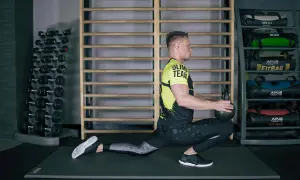 Mężczyzna wykonuje ćwiczenia mobilizacyjne z kettlebell, pogłębiając wykrok. W tle stojaki z hantlami i fitbagami.