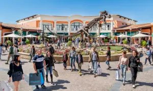 Un T-Rex scheletrico domina il centro del campo rom Castel Romano, circondato da negozi e gente che fa shopping.